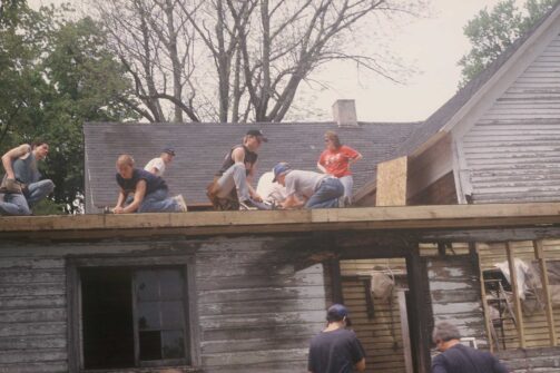 A group of people working together on the roof of an old wooden house, doing repairs or construction outside on a sunny day.