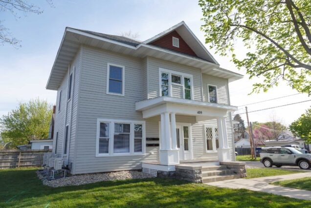 Light gray two-story house with a small balcony, big windows, and a covered front porch, sitting on a green lawn under a bright sky.