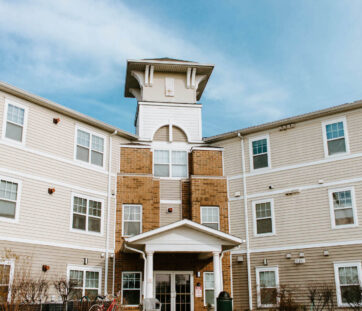 A three-story apartment building with tan siding, white trim, and a small tower on top, under a bright blue sky.