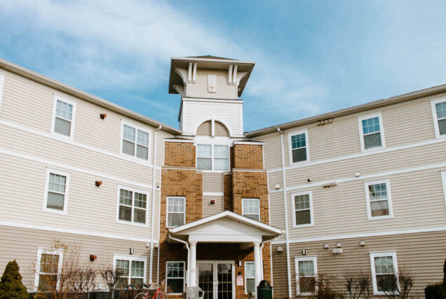 A three-story apartment building with tan siding, white trim, and a small tower on top, under a bright blue sky.