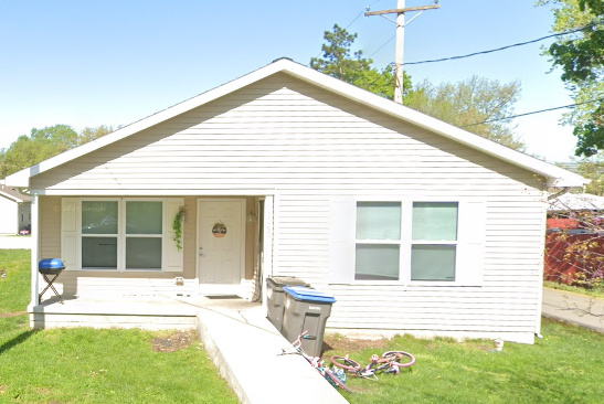 A small white house with a slanted roof, front porch, two trash bins, and a kid’s bike in the yard on a sunny day.
