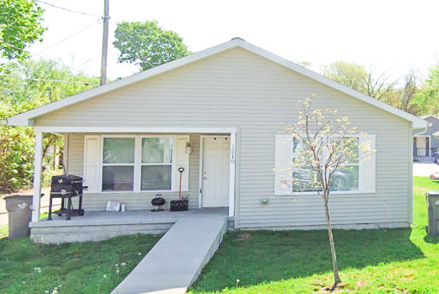 Simple one-story house with a front porch, a small grill, and a single tree in the grassy yard. Bright, sunny day with green trees in the background.