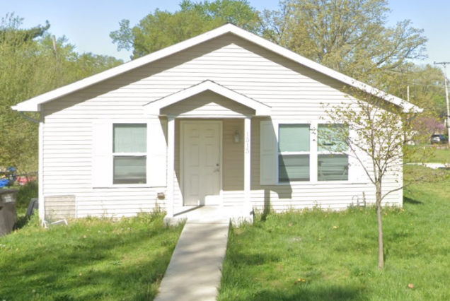 Small white house with a front porch, two windows, a tree in the yard, and a concrete walkway leading to the door.