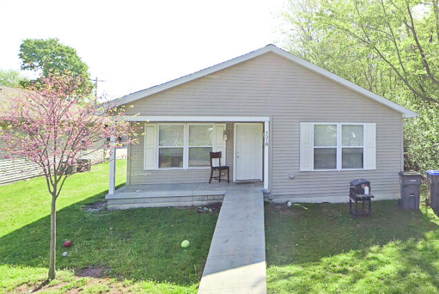 A simple, single-story house with beige siding, a small front porch, a chair, green lawn, some scattered toys, and a tree in the yard.
