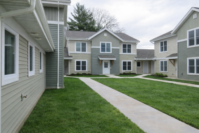 Modern apartment buildings with light-colored siding, green grass, and sidewalks connecting entrances on a cloudy day.
