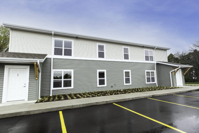 Two-story apartment building with green siding, white trim, and several windows. The parking lot in front has freshly painted yellow lines. Sky is partly cloudy.