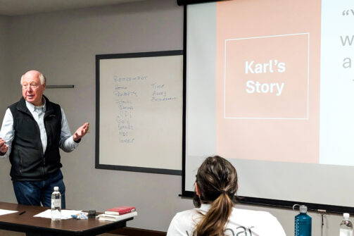 A man gives a casual presentation called "Karl's Story" in a classroom, with a whiteboard listing investment topics beside him.