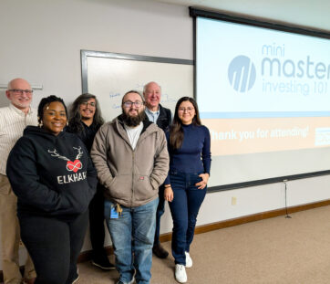 A group of six people smiling in a classroom with a presentation screen that says "mini master investing 101" and "thank you for attending!"