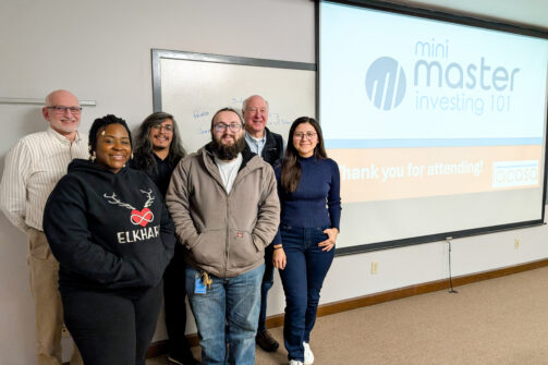 A group of six people smiling in a classroom with a presentation screen that says "mini master investing 101" and "thank you for attending!"