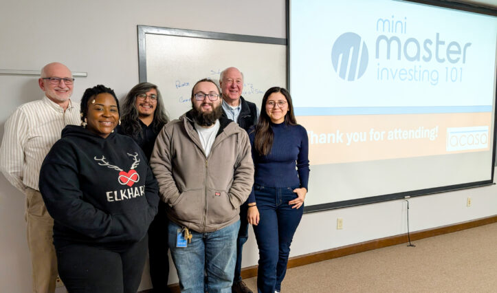 A group of six people smiling in a classroom with a presentation screen that says "mini master investing 101" and "thank you for attending!"