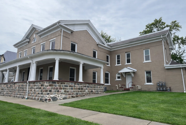 Big old tan house with white trim, a wraparound porch, and stone foundation, sitting on a neat green lawn under a cloudy sky.