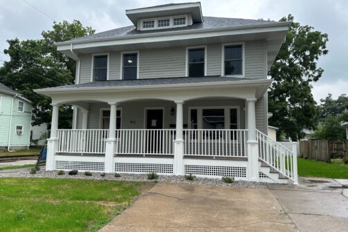 A two-story gray house with a large front porch, white railings, and a small front yard on a cloudy day.
