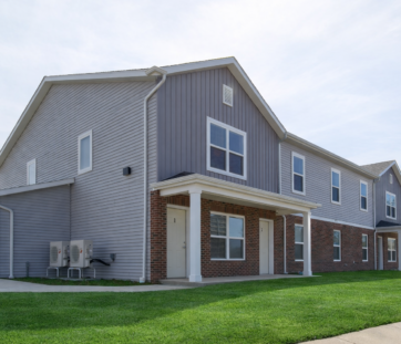 Gray two-story apartment building with brick accents, white trim, and a green lawn out front, on a clear day.