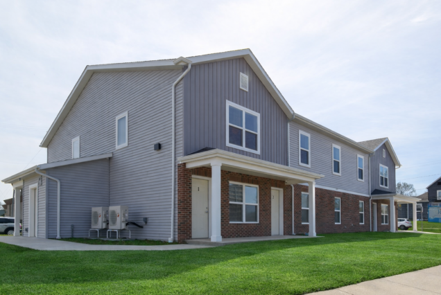 Gray two-story apartment building with brick accents, white trim, and a green lawn out front, on a clear day.