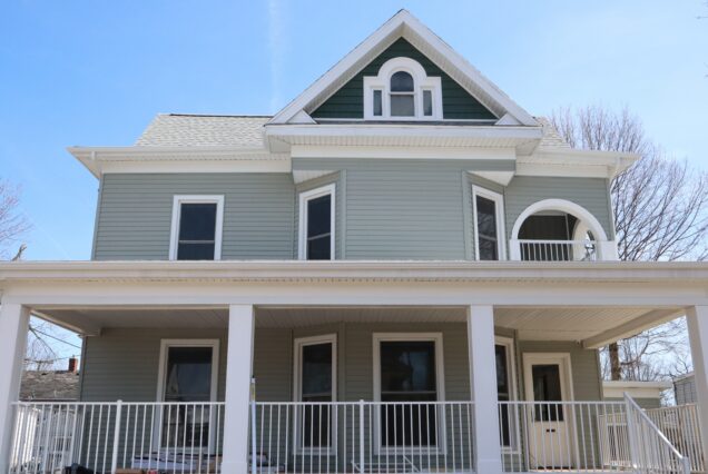 A light gray two-story house with a big porch, white trim, green accents, and a pointed roof, under a clear blue sky.