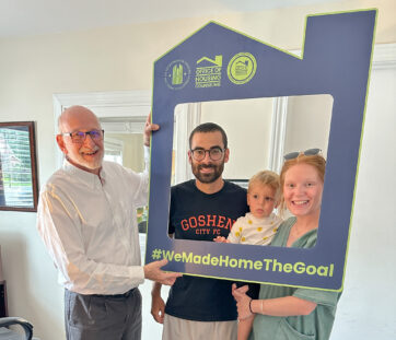 Three adults and a toddler smiling, holding a large blue house-shaped frame that says #WeMadeHomeTheGoal inside a cozy room.