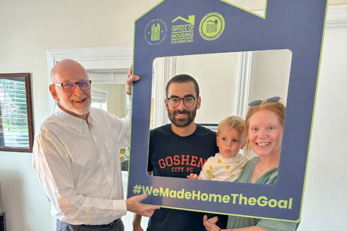 Three adults and a toddler smiling, holding a large blue house-shaped frame that says #WeMadeHomeTheGoal inside a cozy room.