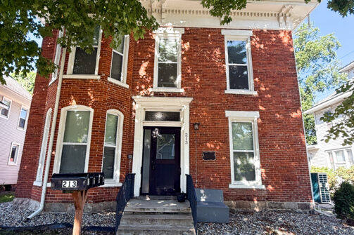 A red brick house with tall windows, white trim, and a black door, shaded by a big tree. Mailboxes labeled 213 are out front.