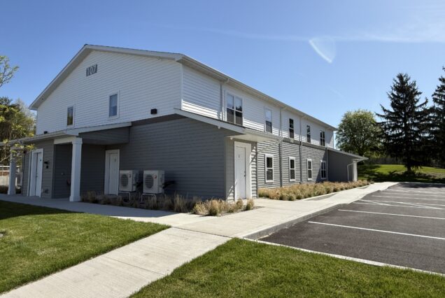 Modern two-story apartment building with gray and white siding, empty parking spaces, green grass, and trees in the background on a sunny day.