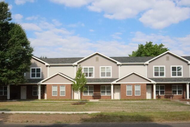 A row of modern townhouses with brick and siding, a couple of trees, and a blue sky with clouds overhead.