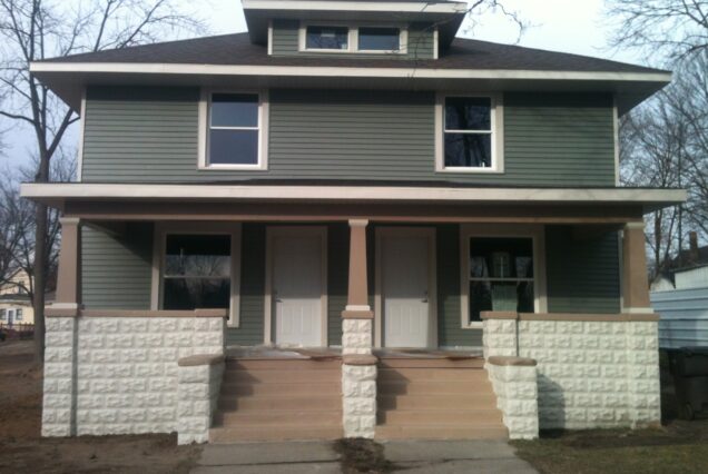 A duplex house with green siding, white trim, two front doors, and a shared porch, set on a quiet street with trees and bare ground.