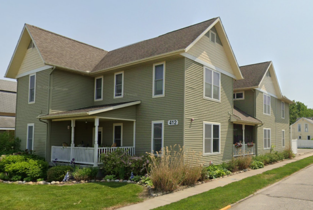 A two-story green house with a porch, white trim, and some plants in the yard, located on a street corner.