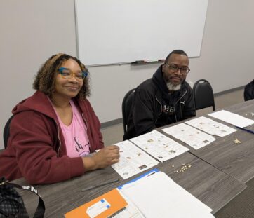 Two people sitting at a table with papers, seeds, and notebooks in a classroom setting, both smiling and looking at the camera.