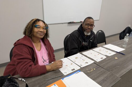 Two people sitting at a table with papers, seeds, and notebooks in a classroom setting, both smiling and looking at the camera.