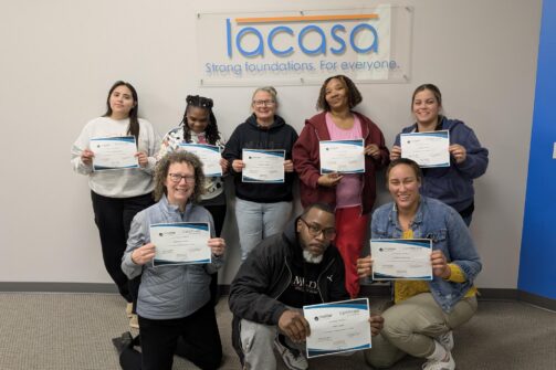 A group of eight people smiling and holding certificates in front of a wall with a "lacasa" sign.