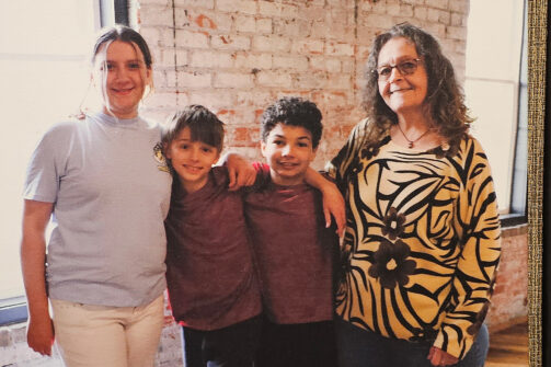 Four people smiling, standing close together indoors by a brick wall and window, looking happy and casual.