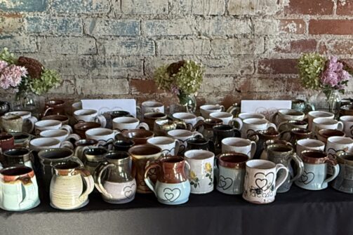 A table with lots of handmade mugs, flowers, and a "lacasa" banner with the slogan "Strong foundations. For everyone." in a rustic room.