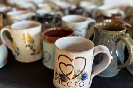 A bunch of handmade ceramic mugs sitting on a black tablecloth, one featured in front says "La Casa" with a heart and floral design.