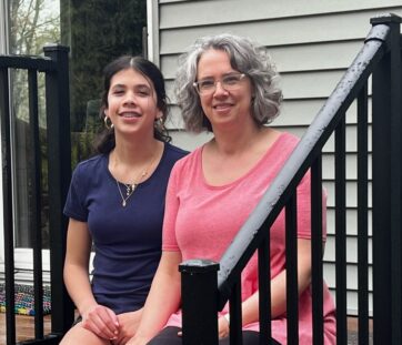 Two women sitting together on outdoor steps, smiling casually. One wears a navy t-shirt, the other wears a pink one. There are black railings and a house in the background.