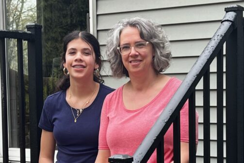 Two women sitting together on outdoor steps, smiling casually. One wears a navy t-shirt, the other wears a pink one. There are black railings and a house in the background.