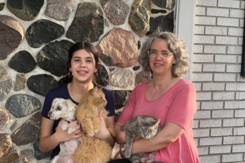 Two women sitting by a stone wall, smiling, each holding pets—one with a dog and a cat, the other with a gray cat.