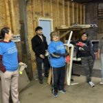 A group of people in casual clothes, some wearing blue volunteer shirts, stand and chat inside a workshop or storage area with wooden walls.
