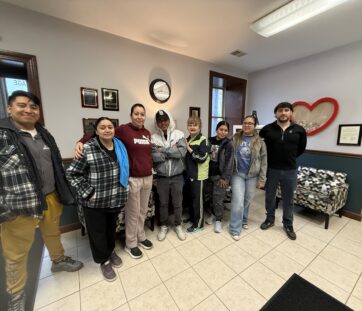 A group of eight people smiling and standing together inside an office with a heart decoration on the wall.