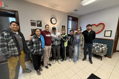 A group of eight people smiling and standing together inside an office with a heart decoration on the wall.