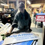 A woman in glasses wipes a Help-A-House sign in a cluttered workshop with Open House signs and various supplies around her.