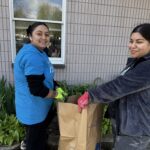Two women smiling while gardening outside, wearing gloves and putting yard waste into a large brown paper bag near a brick building.