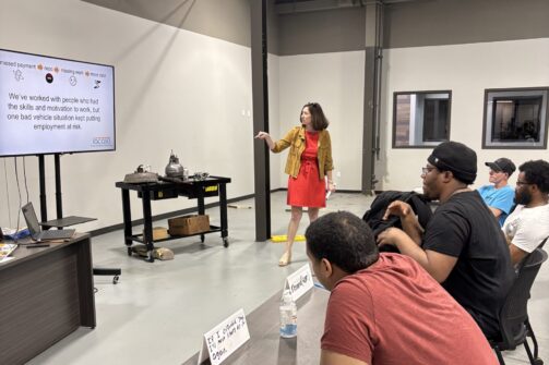 A woman in a red dress presents to a group in a modern classroom, with a presentation slide on a large screen and students sitting at desks.