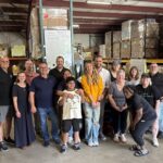 A smiling group of people posing in a warehouse filled with boxes and shelves, looking happy and relaxed, possibly volunteering together.