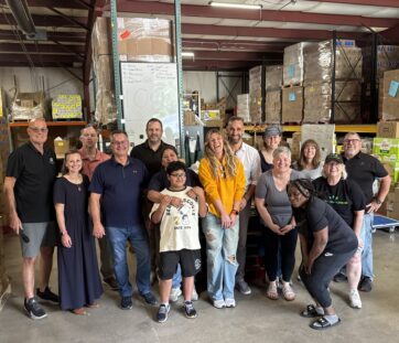 A smiling group of people posing in a warehouse filled with boxes and shelves, looking happy and relaxed, possibly volunteering together.