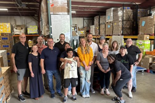 A smiling group of people posing in a warehouse filled with boxes and shelves, looking happy and relaxed, possibly volunteering together.
