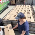 A woman in a blue shirt hands a cardboard box to another person beside a trailer full of boxes, with a "Hunger Ends Here" van nearby.