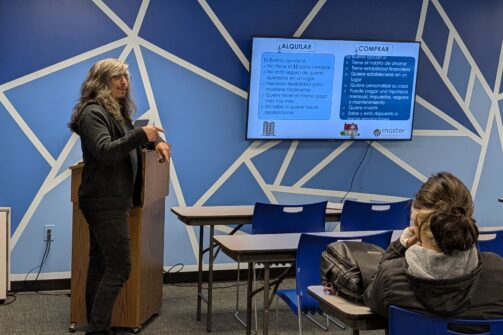 A person is giving a presentation in a classroom with blue geometric walls, while one attendee listens and a screen displays information about renting and buying.
