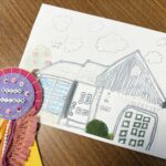 A kid’s drawing of a house with clouds and trees, next to a colorful felt ribbon that says "Dream Builder" on a wooden table.