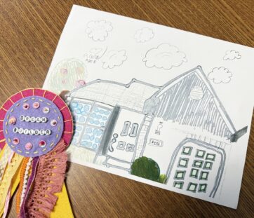 A kid’s drawing of a house with clouds and trees, next to a colorful felt ribbon that says "Dream Builder" on a wooden table.