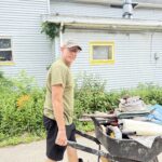 Man in boots and shorts pushing a wheelbarrow full of tools and debris outside a house with gray siding and yellow-trimmed window.
