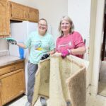 Two women smiling while cleaning a kitchen, one holding a sponge and the other holding up an old piece of carpet.